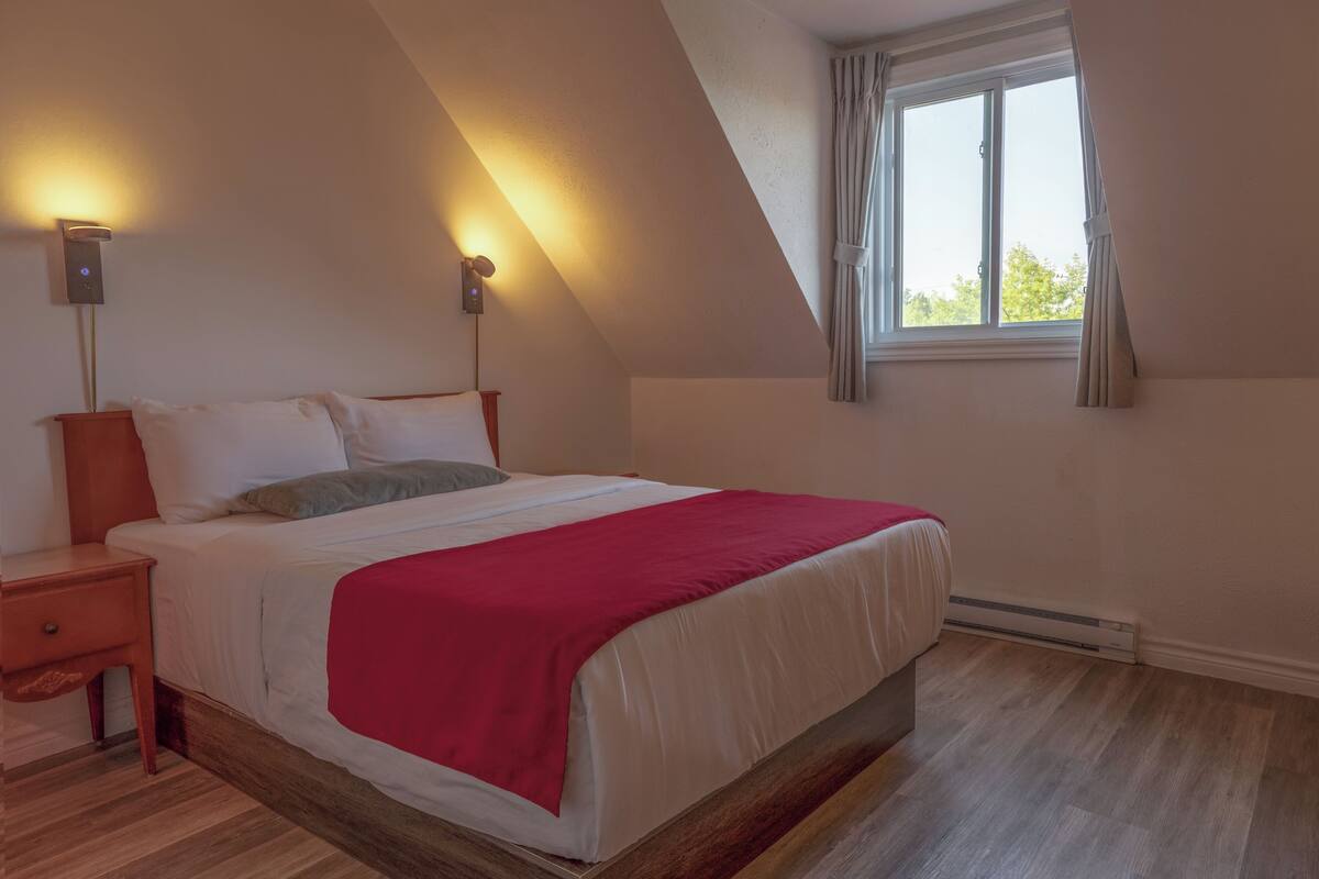 Un dormitorio con cama, mesitas de noche, una colcha roja, una ventana con cortinas e iluminación en la pared.