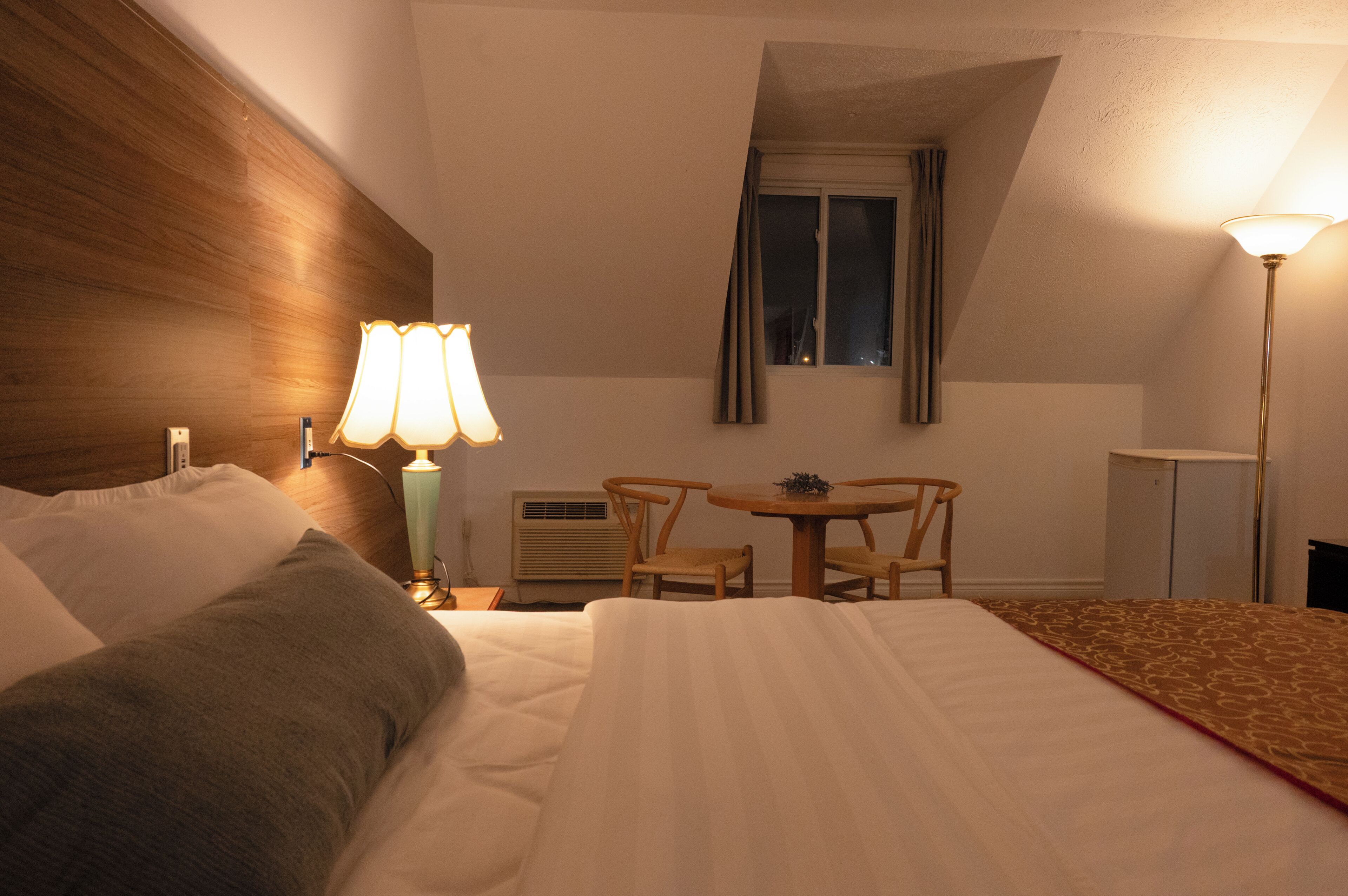 A hotel room with a bed, bedside lamps, a small table with chairs, a window, and an air conditioning unit.