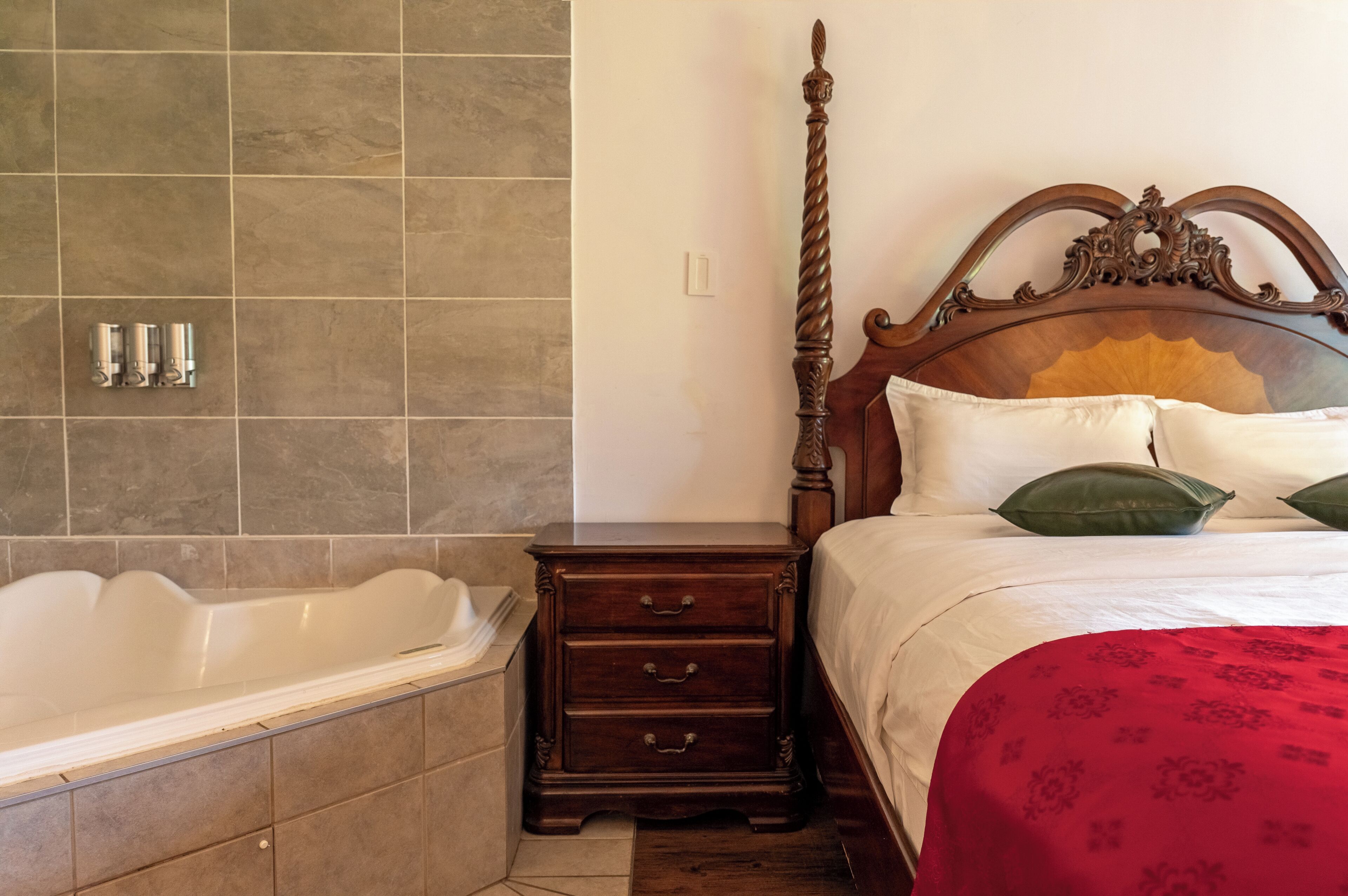 Un dormitorio con una cama con dosel de madera, una mesita de noche con cajones, una bañera blanca y una pared con azulejos.