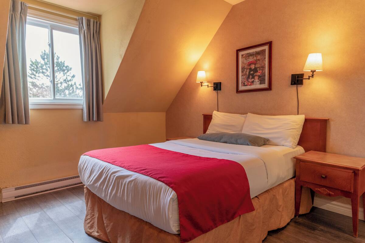 Un dormitorio con cama, mesita de noche, lámpara de pared y ventana con cortinas.