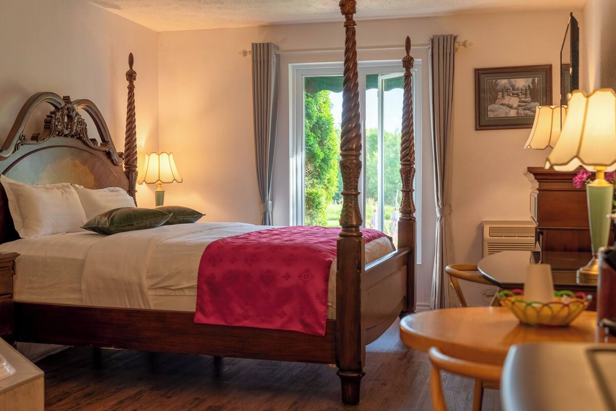 Una cama con dosel con cabecera de madera, una mesita de noche con lámpara, una silla y vistas a la vegetación exterior.