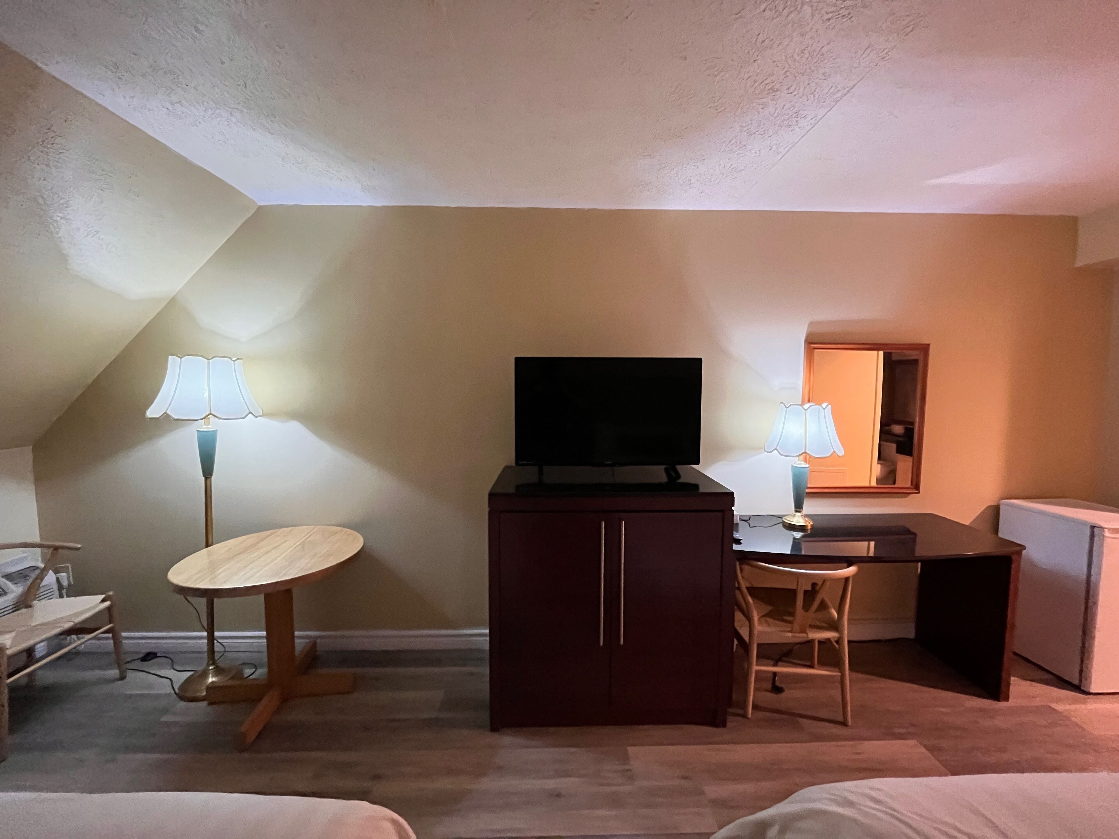A hotel room with a desk, a television, a chair, and a small table with a lamp.