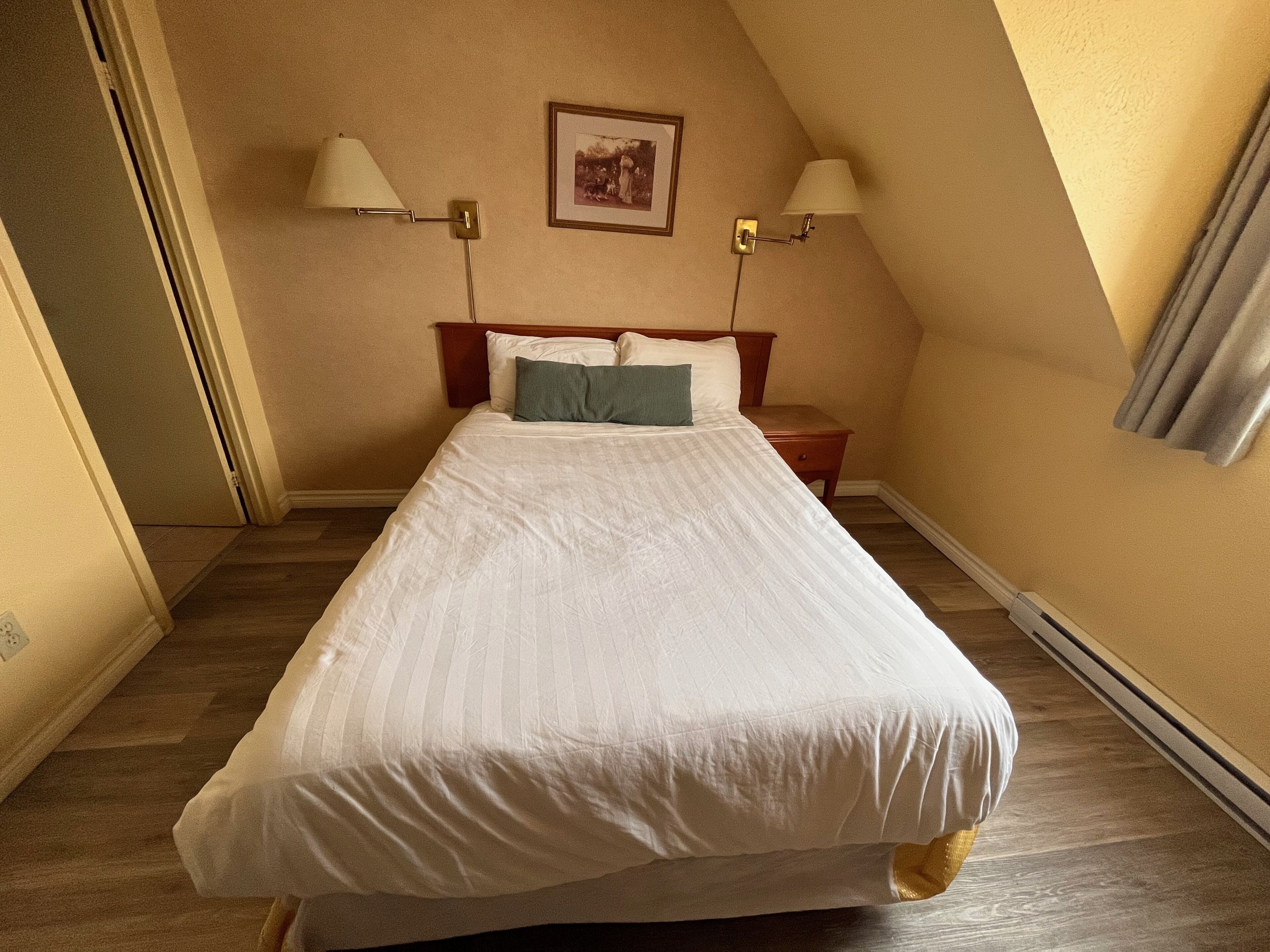 A neatly made bed in a room with a sloped ceiling, two bedside lamps, a framed picture on the wall, and a small bedside table.