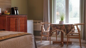 A room with a bed, a table with a floral tablecloth, a chair, a coffee maker, and a window with curtains. - Hotel Cowansville (Cowansville)