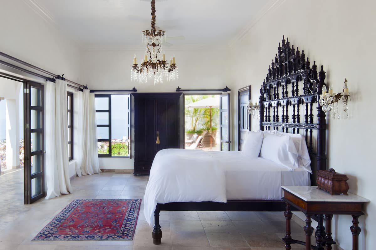 A bedroom with a large bed, a chandelier, a dark wooden headboard, and a view of the outdoors.