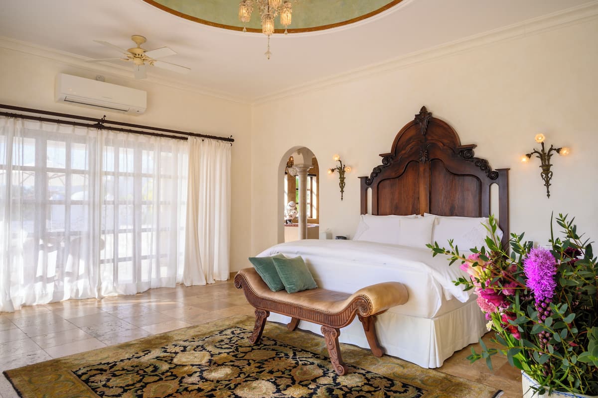 A spacious bedroom with a large bed, a wooden bench, a decorative chandelier, and a potted plant.