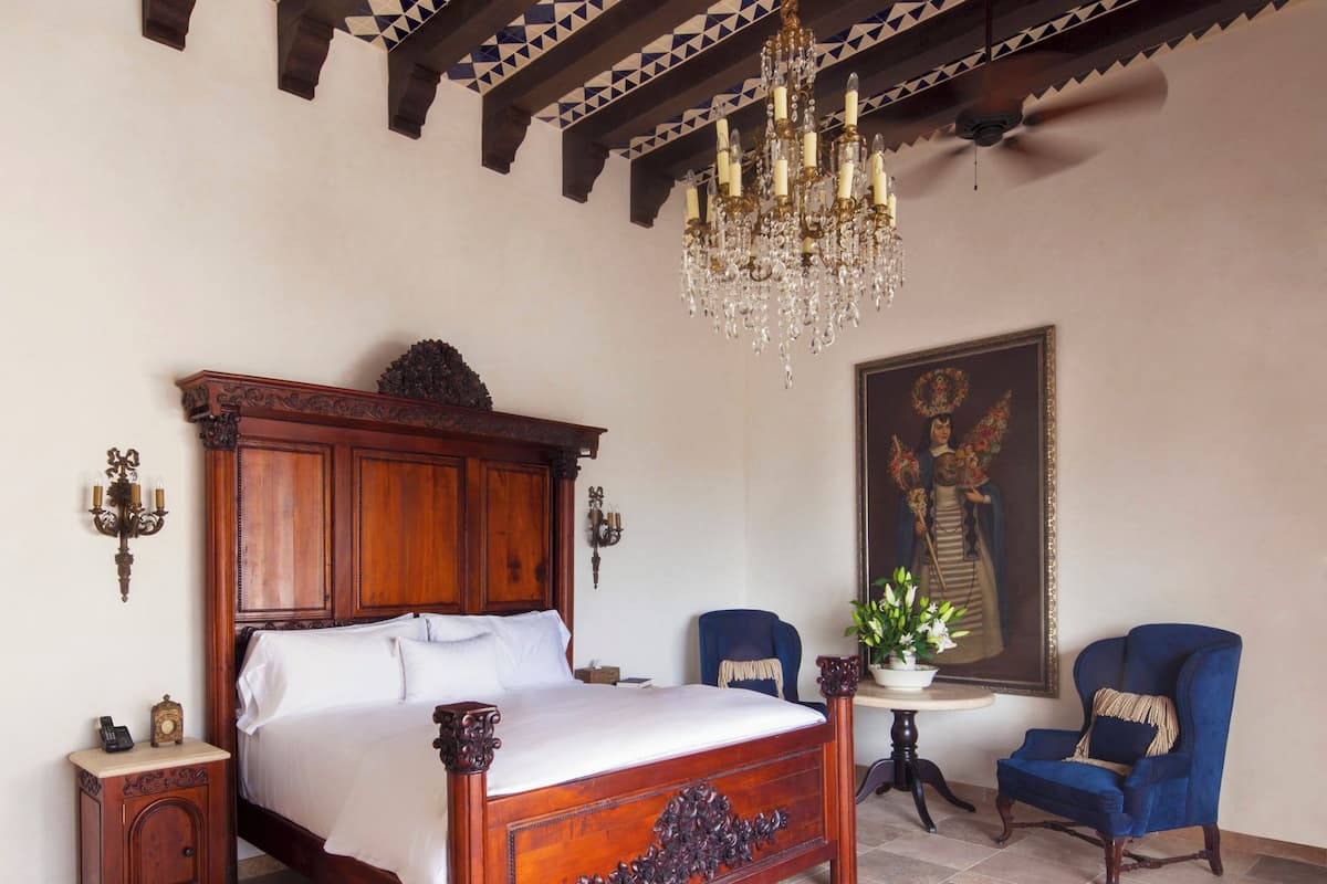 A bedroom with a large wooden bed, a chandelier, a blue armchair, a small table with a plant, and a painting on the wall.