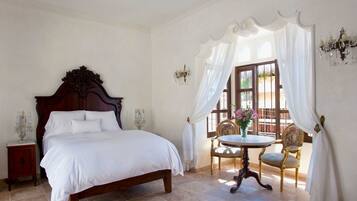A bedroom with a large bed, a round table with chairs, a window with curtains, and a chandelier.