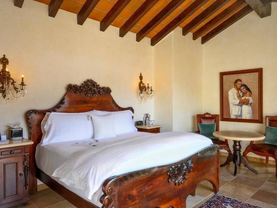 A bedroom with a large wooden bed, two bedside tables, a small round table, two chairs, and a painting on the wall.