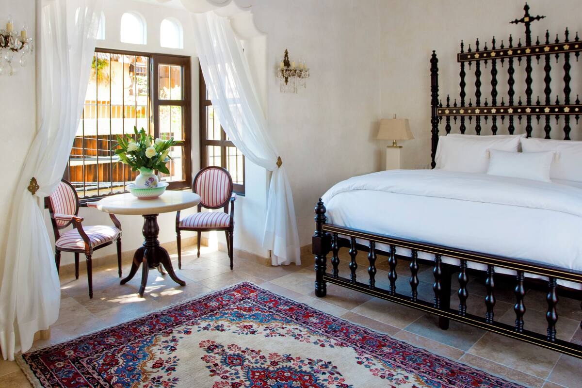 A bedroom with a large bed, a small table with chairs, a decorative rug, and a window with curtains.