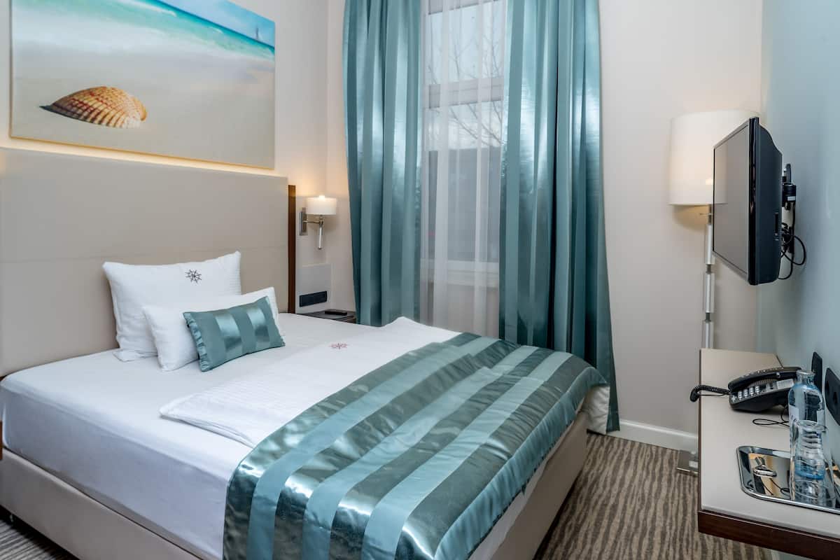 A hotel room with a neatly made bed, a desk with a telephone and a bottle, and a painting of a shell on the wall.