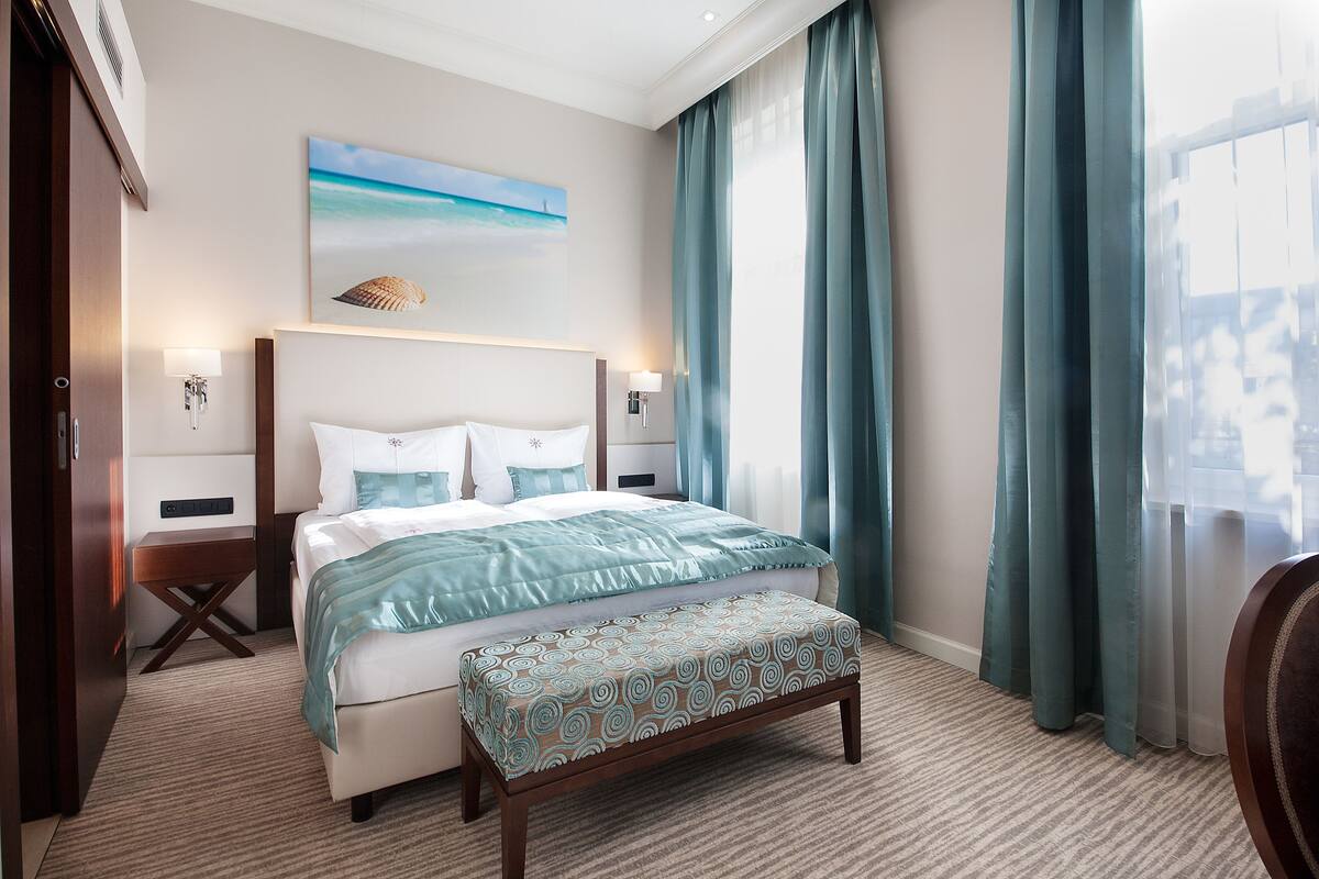 A hotel room with a large bed, a bedside table, a bench, a painting of a beach, and a view of the outdoors through the window.