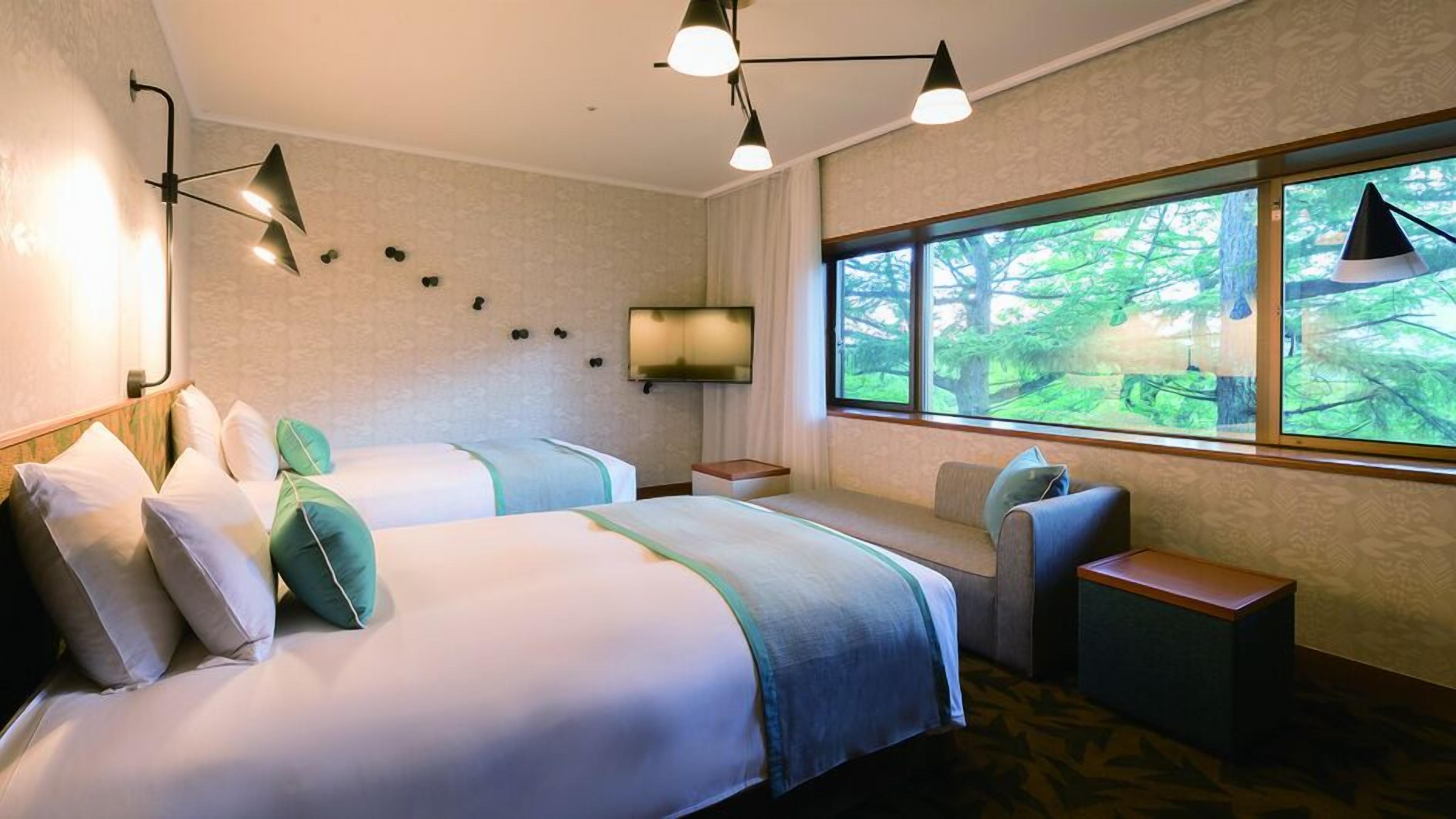 A hotel room with two beds, a TV, a window with a forest view, and a small seating area.