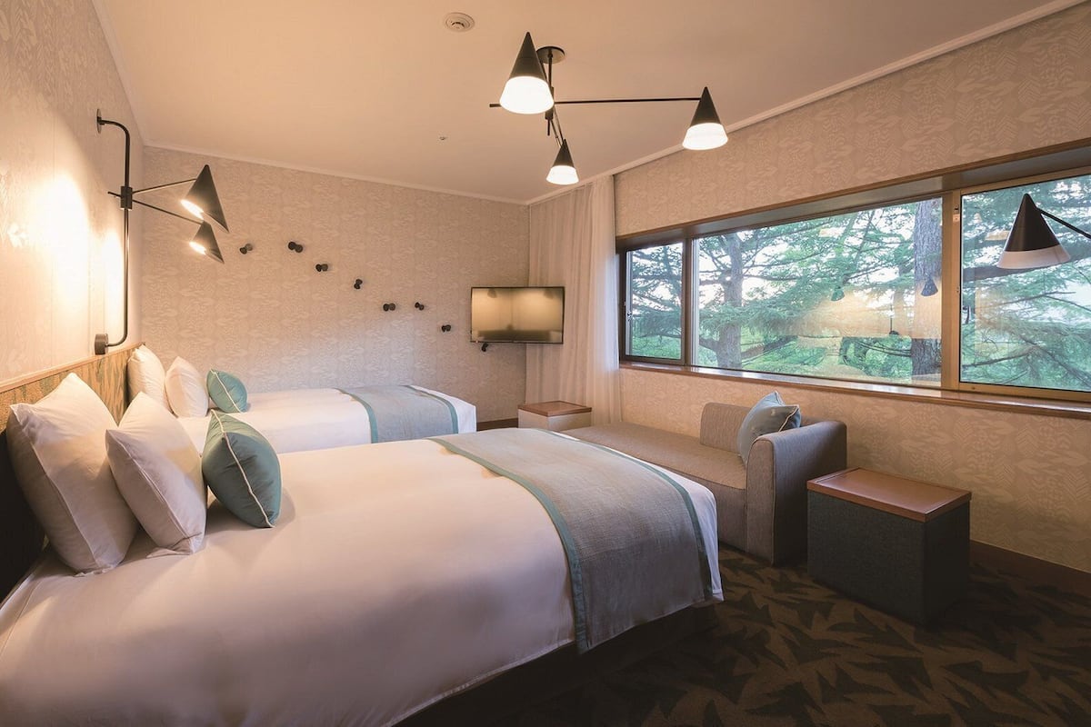 Une chambre d’hôtel avec deux lits, une télévision, une fenêtre donnant sur la forêt et un bureau.