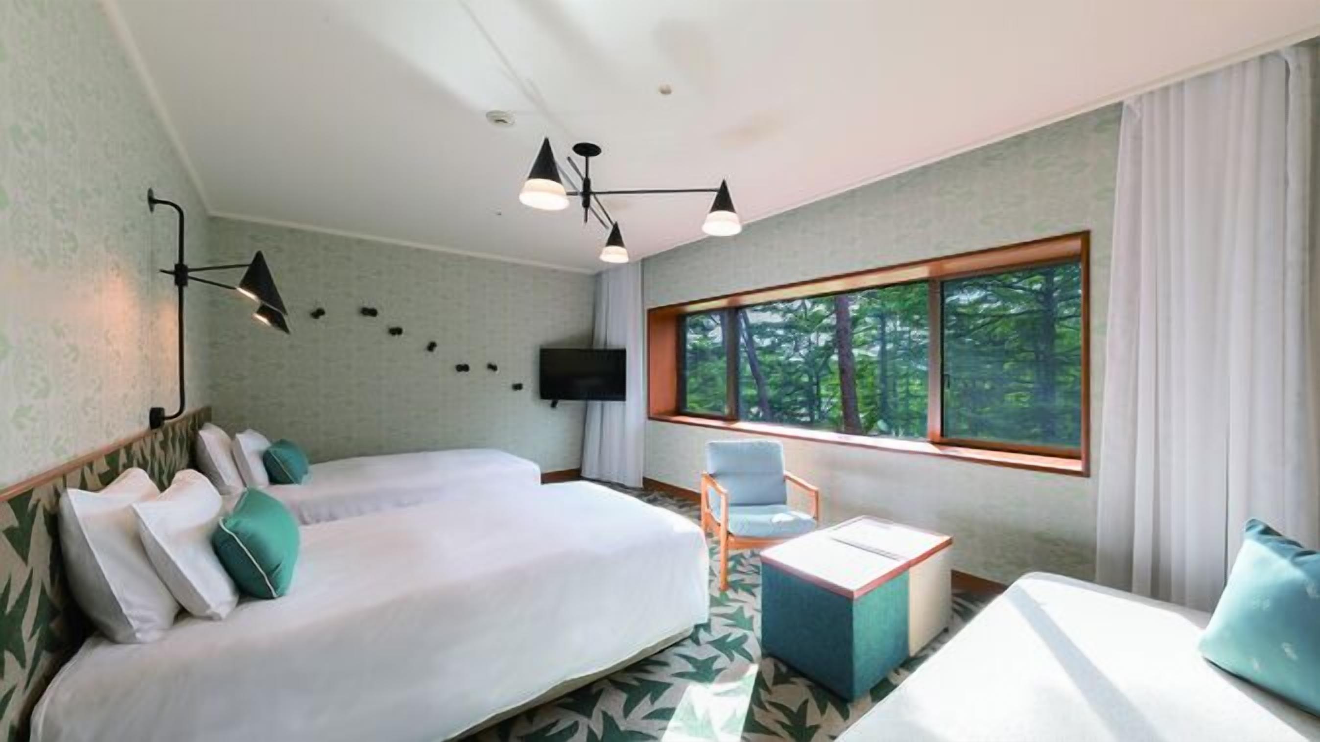 A hotel room with a bed, a chair, a TV, and a window with a forest view.