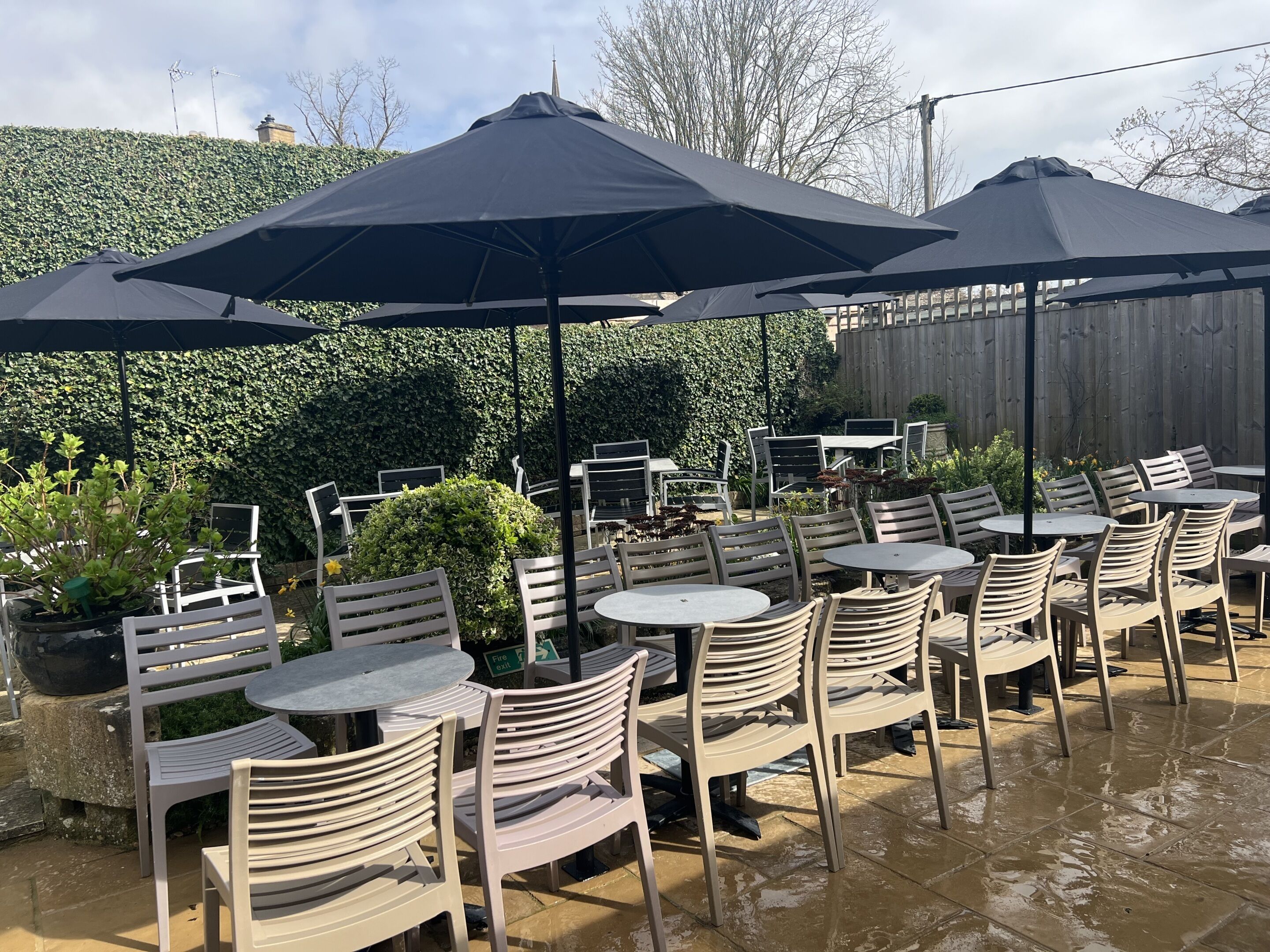 An outdoor patio with round tables and chairs, covered by large umbrellas, and surrounded by greenery.