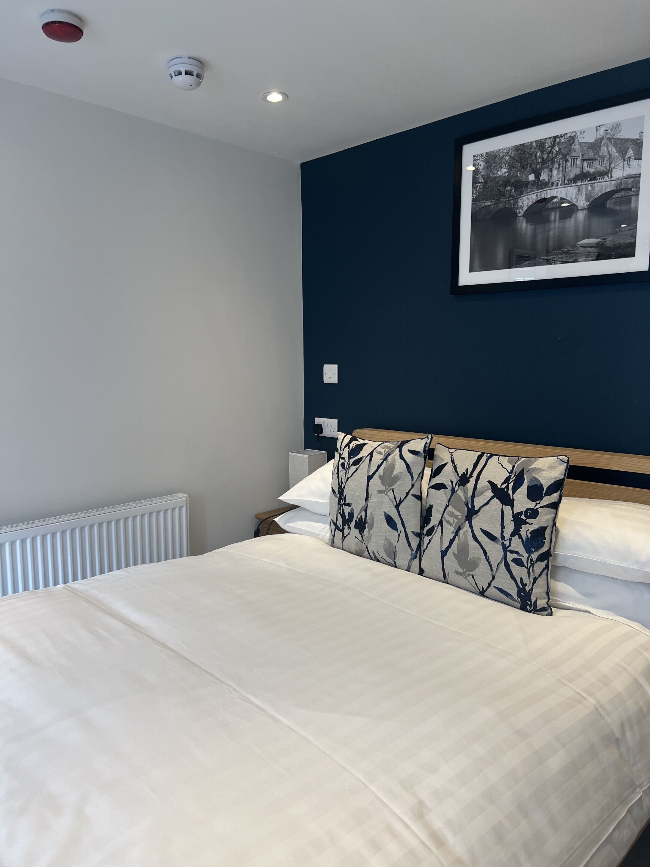 Una cama bien tendida con sábanas blancas y un cojín estampado con motivos florales, una fotografía en blanco y negro enmarcada en la pared y un radiador blanco.