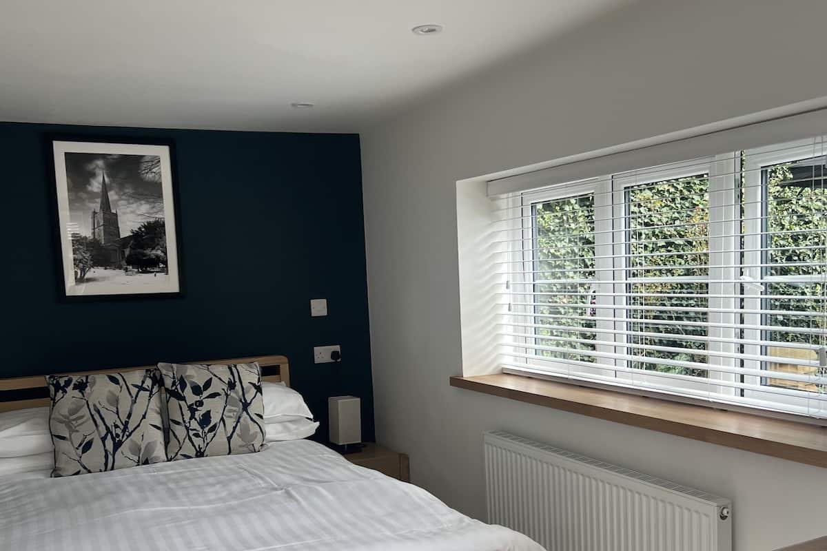 A bedroom with a bed, a bedside table, a window with blinds, a radiator, and a framed picture on the wall.