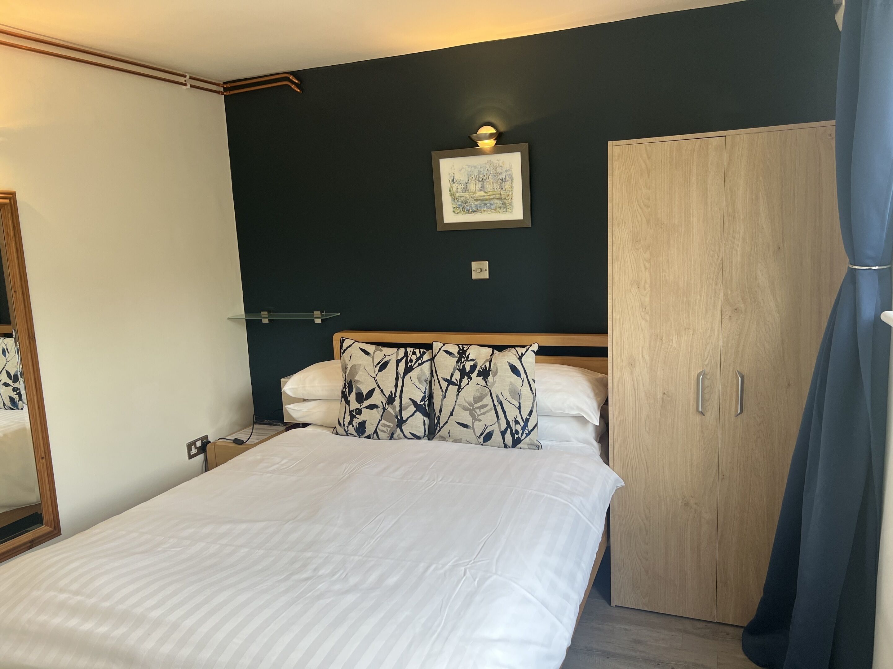 A bedroom with a bed, a wardrobe, a bedside table, a lamp, and a framed picture on the wall.