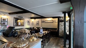 En restaurang med träbord och stolar, en disk med bakverk och bröd, inramade tavlor på väggarna och en stenmur i bakgrunden.
