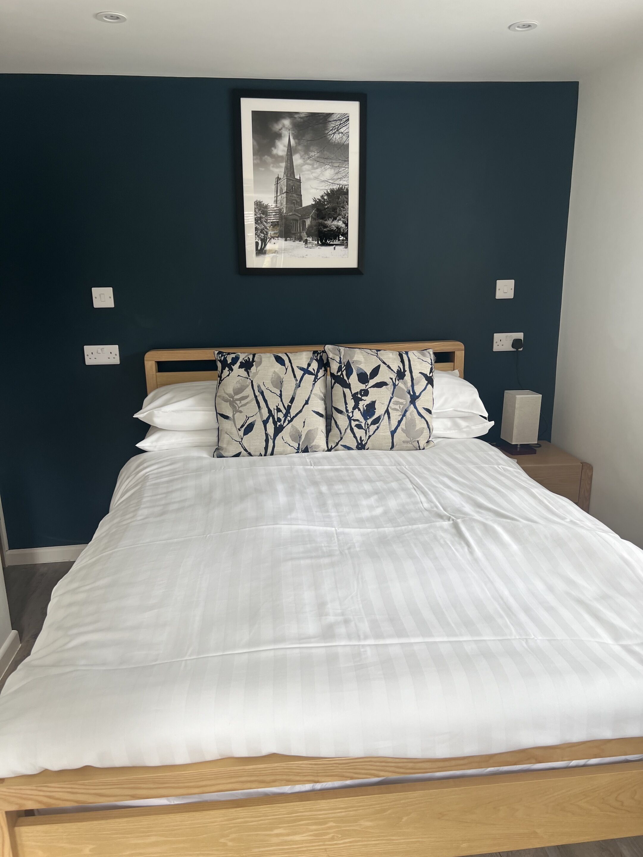 Una cama bien hecha con ropa blanca y dos almohadas decorativas, un cabecero de madera y un cuadro con una vista de un edificio en la pared.