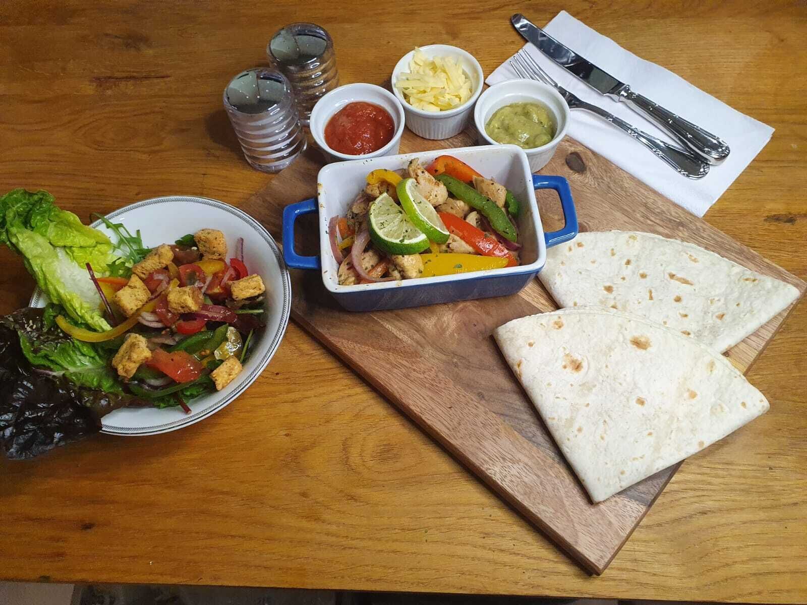 Une table en bois sur laquelle est dressé un repas composé d’une salade, d’un plat de légumes grillés et de poulet, de deux tortillas et de divers condiments.