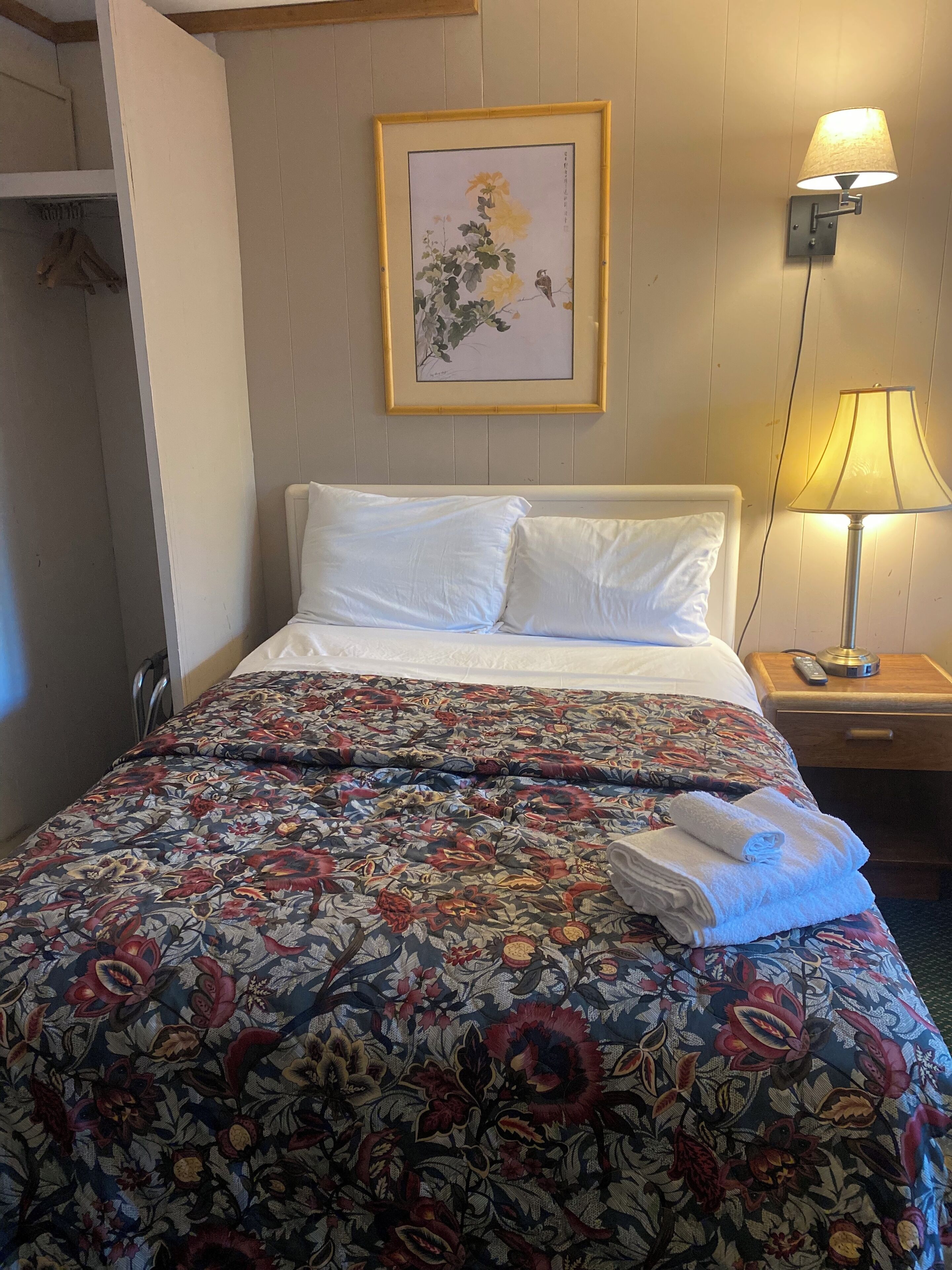 A hotel room with a neatly made bed, a framed floral artwork on the wall, a bedside lamp, and a folded towel on the bed.