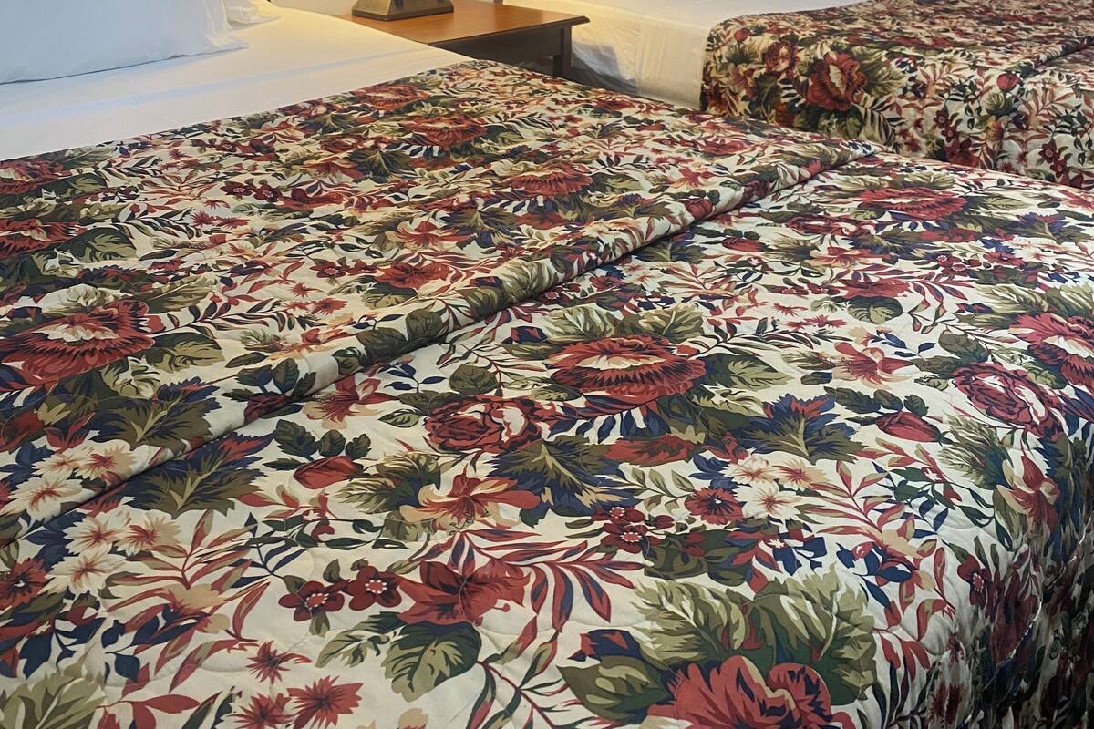 Habitación de hotel con dos camas, cada una con sábanas blancas y colcha estampada con motivos florales. Hay una mesita de noche con una lámpara entre las camas.