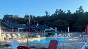A pool area with red and white Adirondack chairs, a fenced swimming pool, and a building with a dark roof in the background.