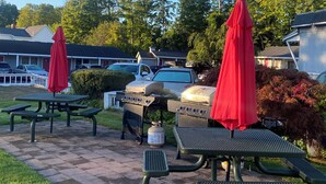 A patio area with picnic tables and barbecue grills, surrounded by trees and a building in the background.