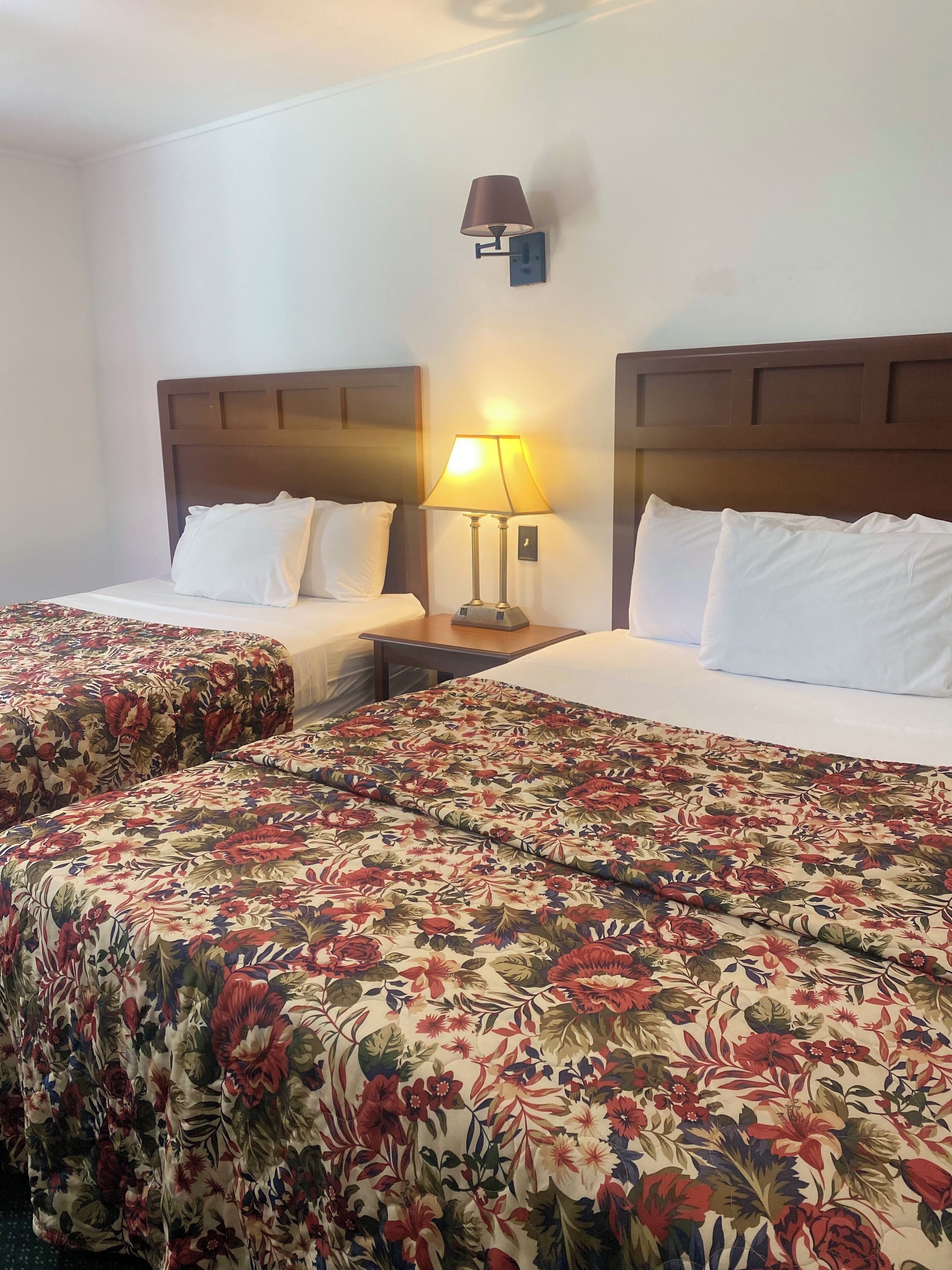 A hotel room with two beds, each with a floral bedspread and white pillows. There is a nightstand with a lamp between the beds.