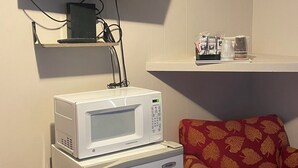 A compact living space with a flat-screen TV mounted on the wall, a white microwave on a small refrigerator, and a shelf with a few items.