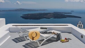A sunlit terrace with white lounge chairs, a straw hat, a wine glass, and a fruit basket.
