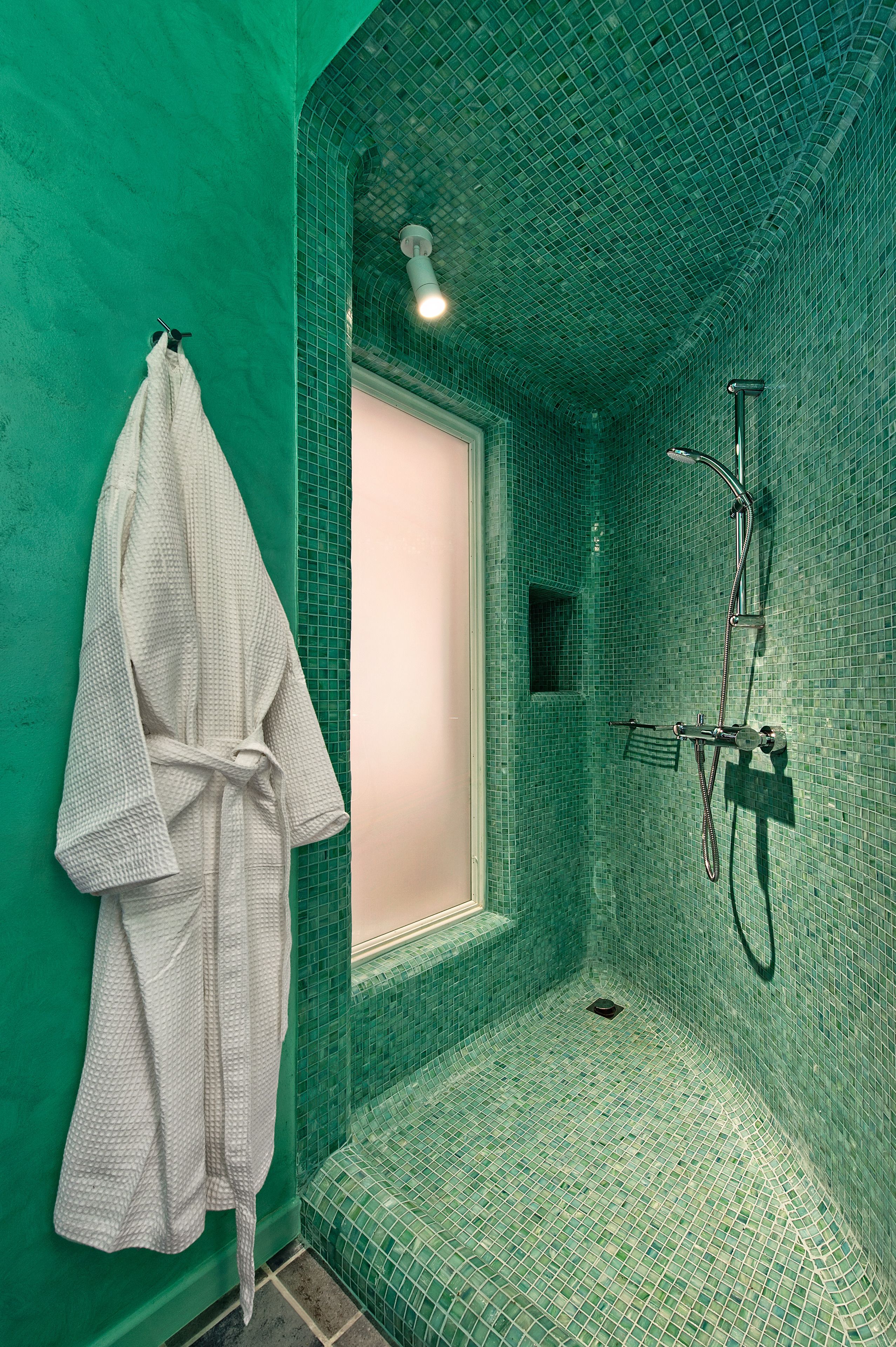 A bathroom with a green tiled wall, a white bathrobe hanging on the wall, and a shower area with a glass door.