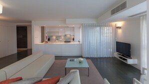 Ein modernes Wohnzimmer mit einer Couch, einem Couchtisch und einem Fernseher. Im Hintergrund ist eine Küche zu sehen.