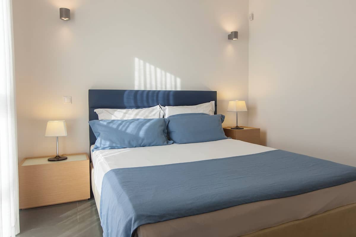 Een net opgemaakt bed met een blauw-witte spreideken, twee nachtkastjes met lampen en een modern hoofdbord.