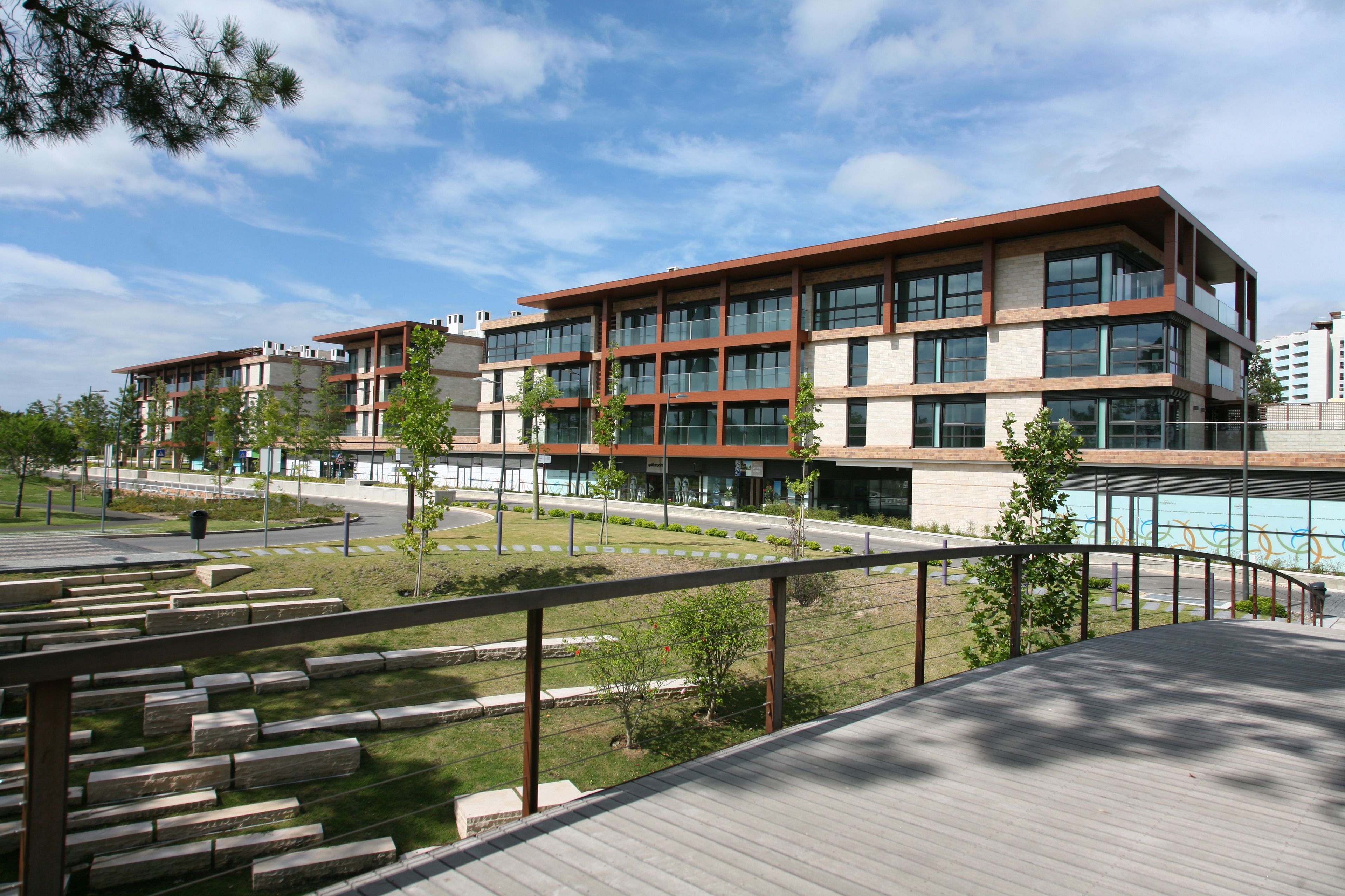 Un edifici modern amb múltiples plantes, grans finestres i un espai verd envoltant amb bancs.