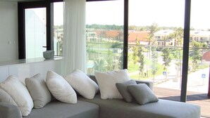 Ein modernes Wohnzimmer mit einer grauen Eckcouch, weißen und grauen Kissen, einem weißen Couchtisch und großen Fenstern mit Blick auf eine Wohngegend mit Häusern und viel Grün.