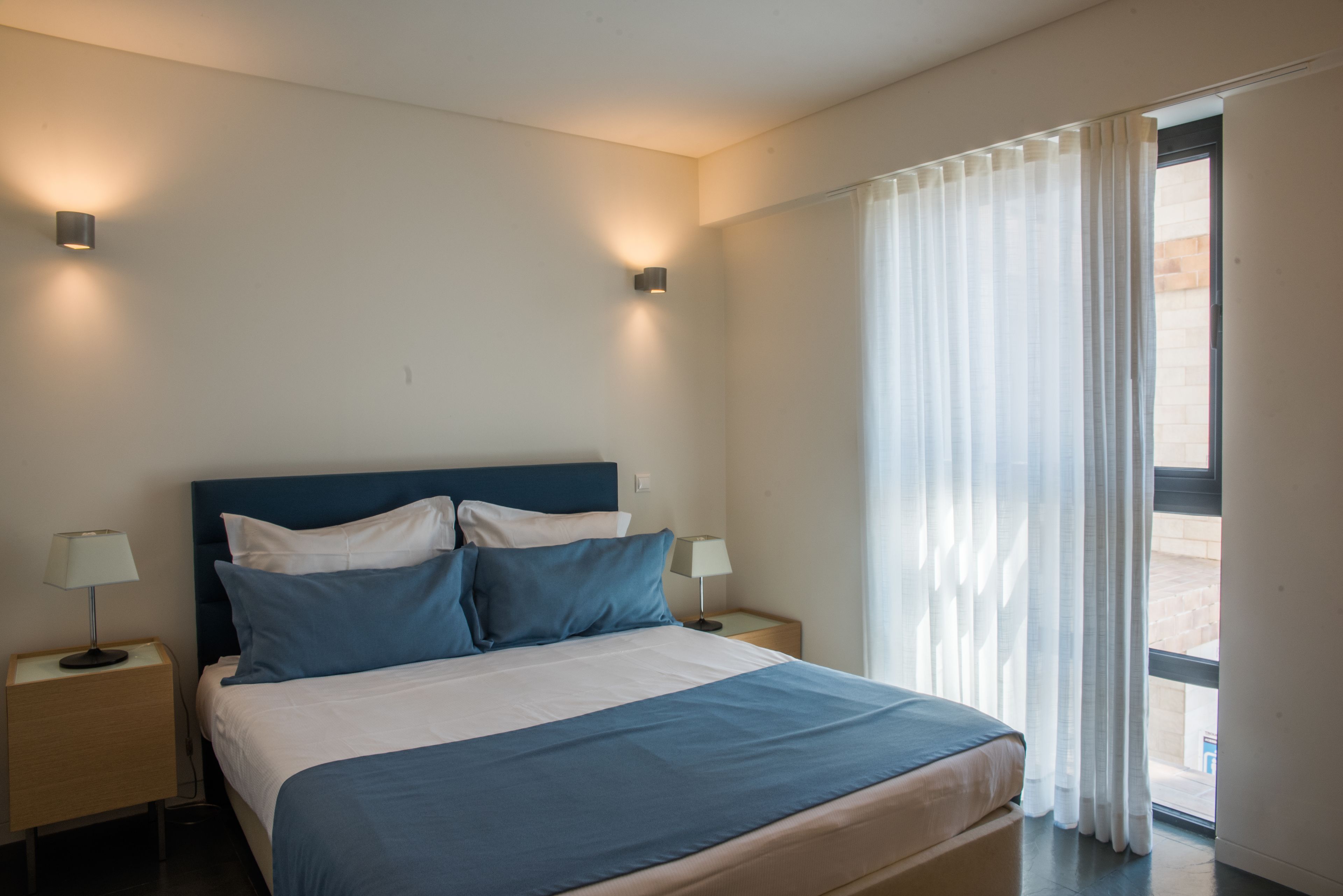 Quarto de hotel com uma cama, duas mesas de cabeceira com abajures, uma janela com cortinas translúcidas e vista para o exterior.