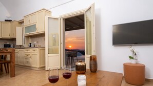 A modern kitchen with wooden cabinets, a dining table with two wine glasses, and a view of a sunset through an open door. - White Orchid Villa (Santorini)
