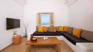 A modern living room with a sectional sofa, a flat-screen TV, a coffee table, and a view of the sea through a window. - White Orchid Villa (Santorini)