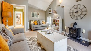 A living room with a grey sofa, a white coffee table, a black wood-burning stove, and a large wall clock. - Broadrock (Chepstow)