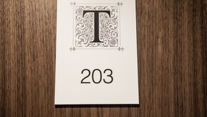 Une carte blanche ornée d’un grand « T » décoratif et du chiffre 203, le tout posé sur une surface en bois.
