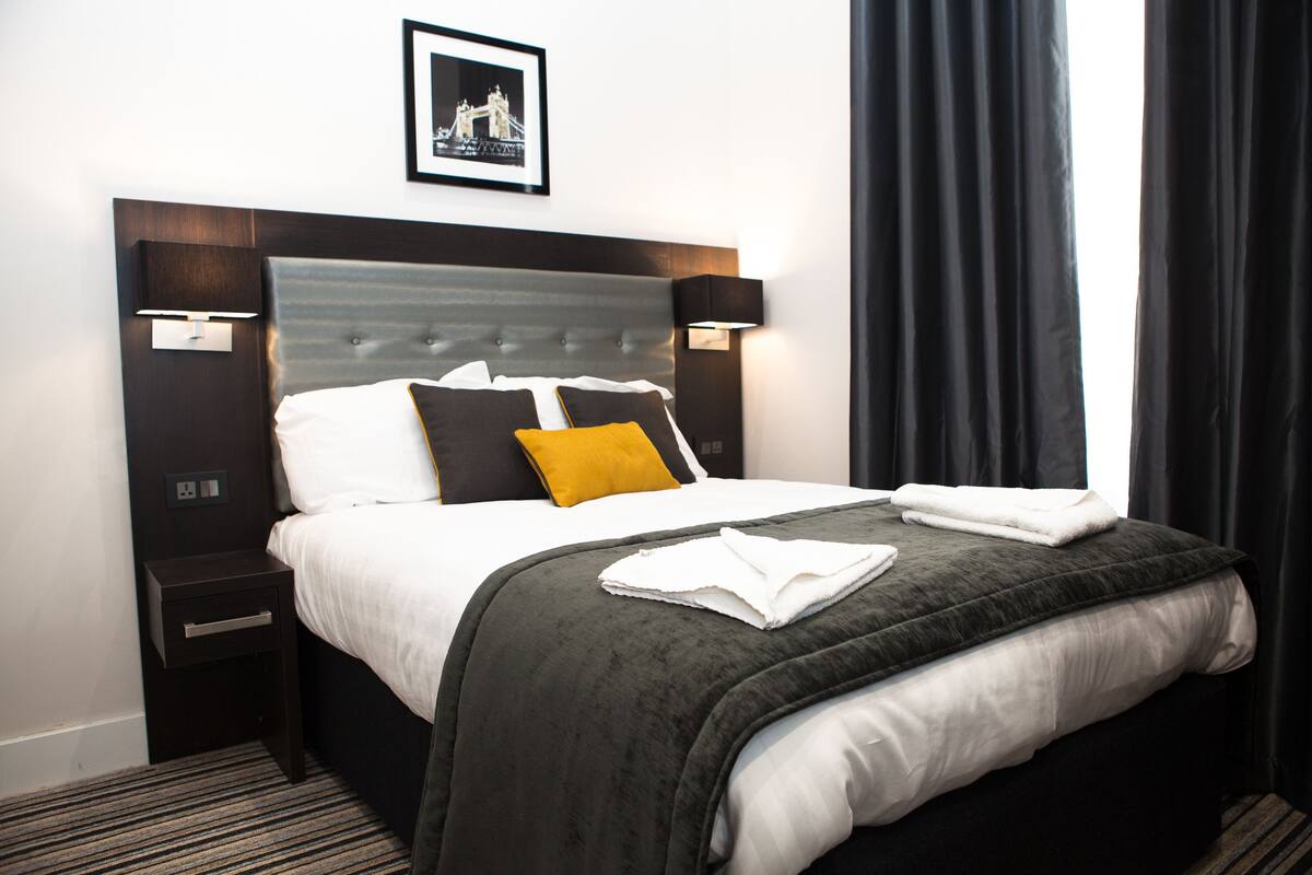 A hotel room with a neatly made bed, a headboard with built-in lighting, a framed picture on the wall, and a window with dark curtains.