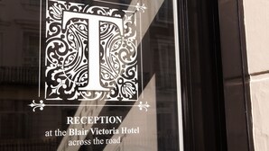 Une enseigne indiquant la réception de l’hôtel Blair Victoria, avec une grande lettre décorative « T » et le texte : « RÉCEPTION à l’hôtel Blair Victoria, de l’autre côté de la rue ».