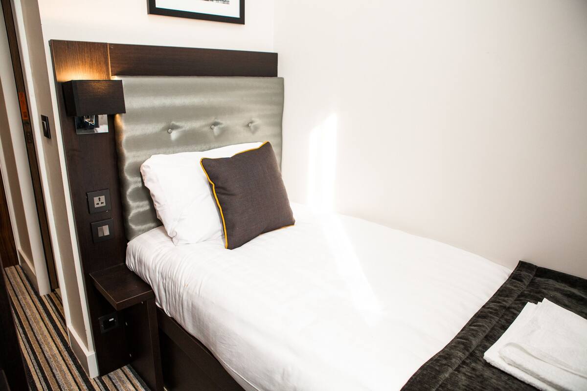A neatly made bed with white linens and two pillows, a bedside table with a lamp, and a framed picture on the wall.