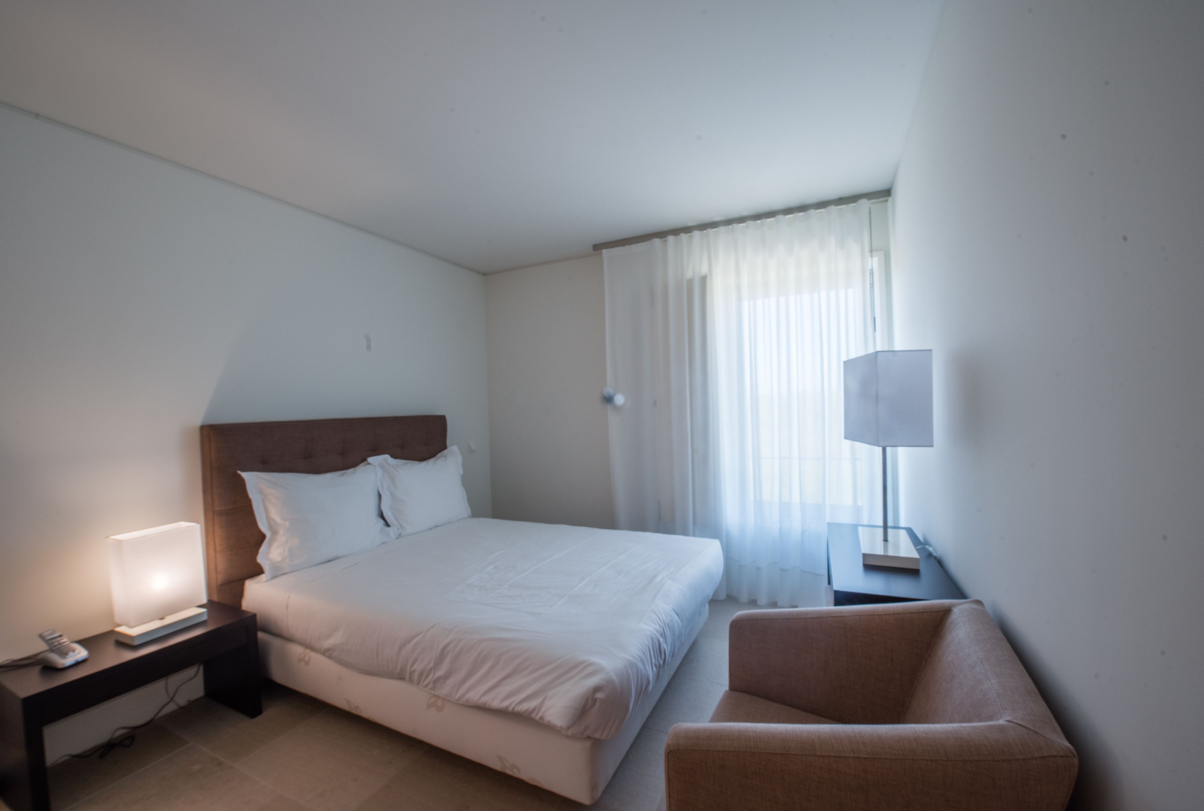A hotel room with a bed, a nightstand with a lamp, a chair, and a window with curtains.