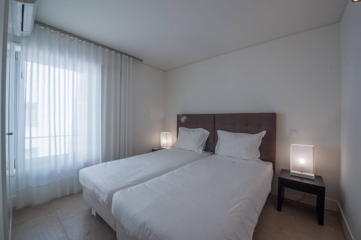 Habitación de hotel con cama doble, dos mesitas de noche con lámparas, cabecero y ventana con cortinas translúcidas.