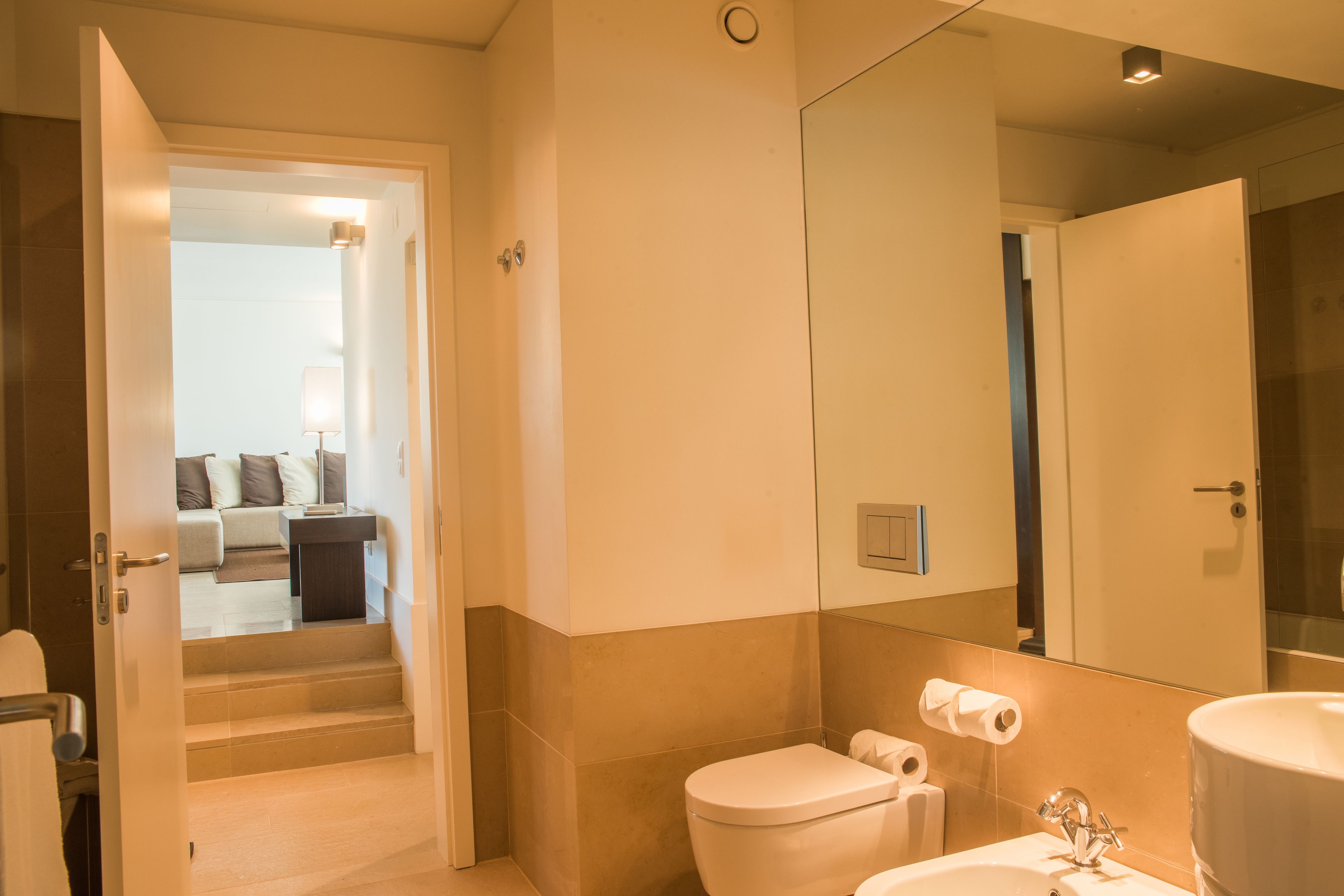 A modern bathroom with a toilet, sink, and mirror. There is a staircase leading to a living area with a sofa and lamp.