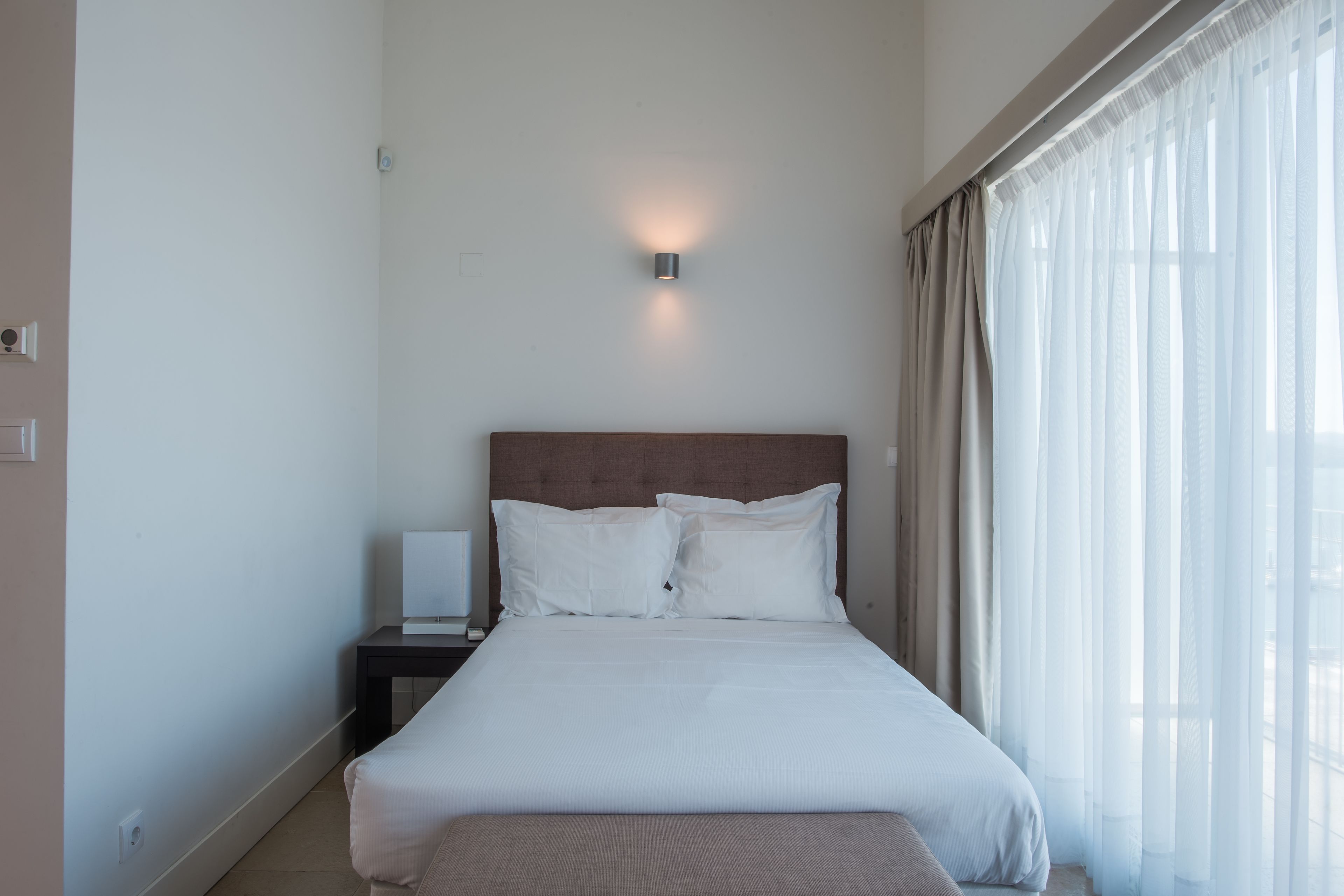 A neatly made bed with white linens, a bedside table with a lamp, and sheer curtains on a window.